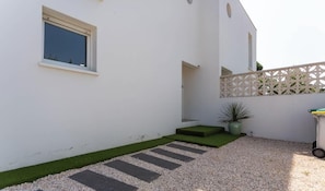 A white building with a window, a small green area, and a gravel path. - Villa 8 pers climatisée avec terrasse proche mer (Sainte-Marie-la-Mer)