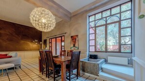 A dining area with a wooden table and chairs, a chandelier, a large window with a view, and a white sofa. - Villa avec piscine 8 pers proche Perpignan (Saleilles)