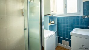 Une salle de bain avec une douche à parois de verre, un lave-linge blanc, des murs recouverts de carreaux bleus et une fenêtre avec des stores.