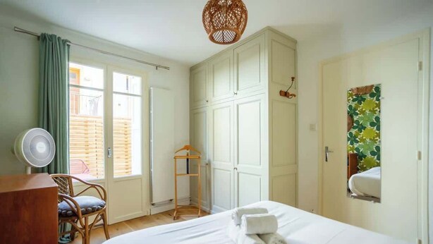 Une chambre à coucher avec un lit, un ventilateur, une chaise, une armoire et une fenêtre avec des rideaux.