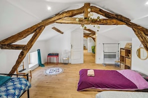A loft bedroom with exposed wooden beams, a bed with a purple bedspread, a small table, a chair, and a fan.