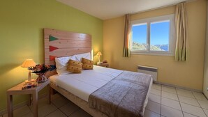 A hotel room with a bed, a nightstand with a lamp, a window with curtains, and a view of the outside. - Mediterranean Sea View Escape for 6 (Cerbère)