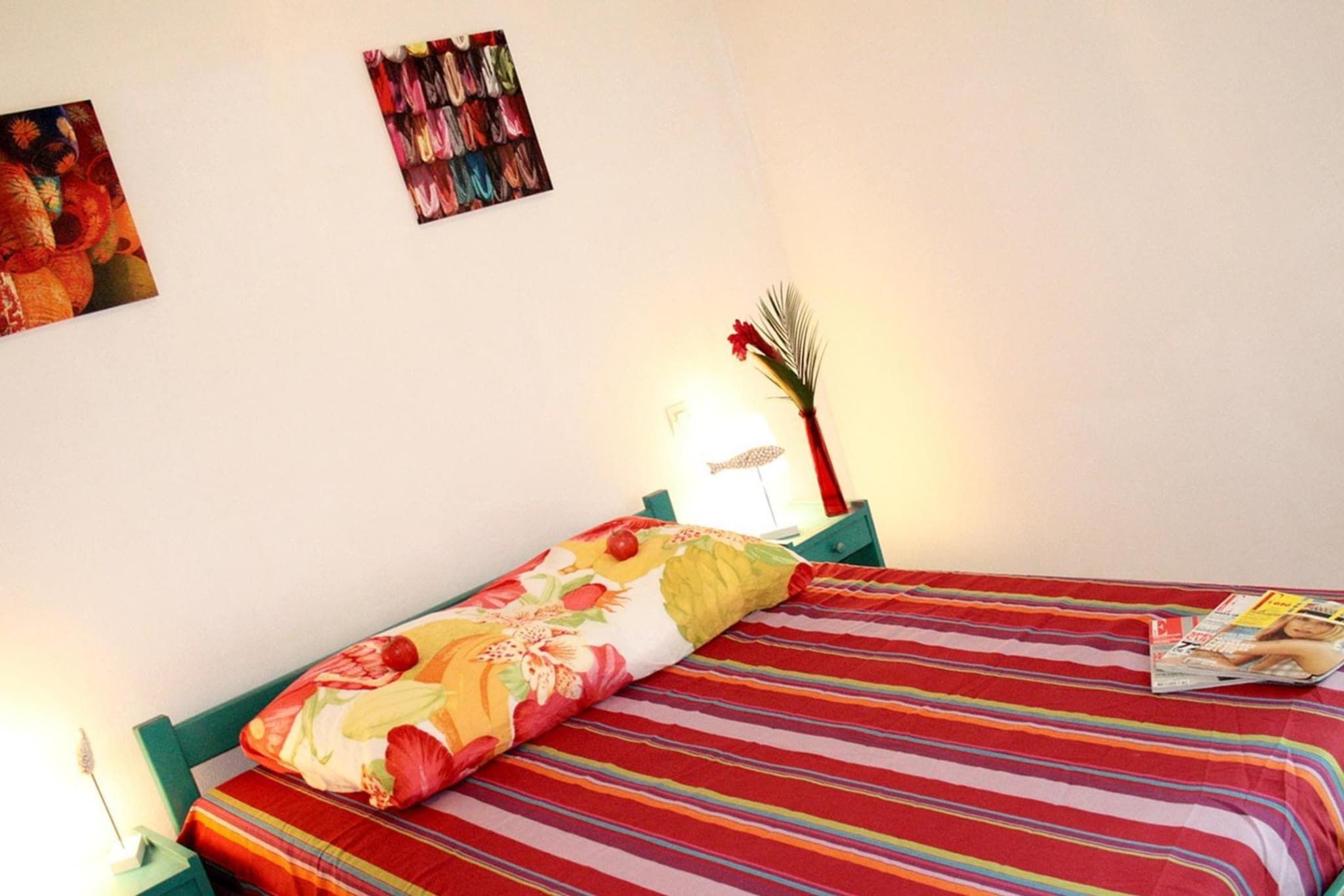 A bed with a striped bedspread and a floral pillow, a magazine on the bed, a lamp, and a wall with a framed picture.