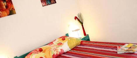 A bed with a striped bedspread and a floral pillow, a magazine on the bed, a lamp, and a wall with a framed picture.