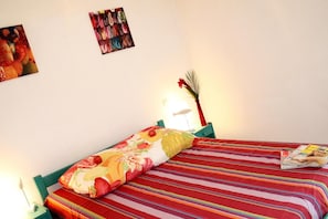 Ein Bett mit gestreifter Bettdecke und einem floralen Kopfkissen, eine Zeitschrift auf dem Bett, eine Lampe und eine Wand mit einem gerahmten Bild.