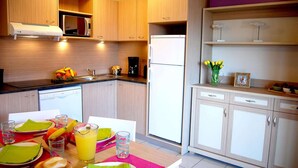 A modern kitchen with wooden cabinets, a microwave, a refrigerator, and a dining table set with plates, glasses, and a fruit bowl. - Spacious Beachside Apartment for 6 (Saint-Cyprien)