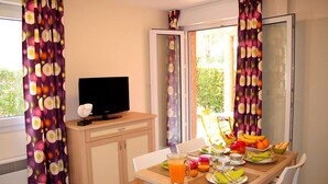 A room with a TV on a wooden cabinet, a dining table with breakfast items, and curtains with a fruit pattern. - Spacious Beachside Apartment for 6 (Saint-Cyprien)