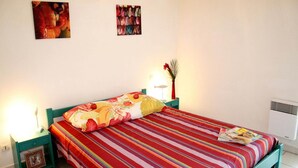 A bed with a striped bedspread, a bedside lamp, a framed picture on the nightstand, and a wall-mounted air conditioner. - Spacious Beachside Apartment for 6 (Saint-Cyprien)