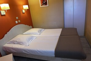 Una cama doble con ropa de cama blanca y marrón, dos lámparas fijadas a la pared, una pequeña estatua y una puerta cerrada.