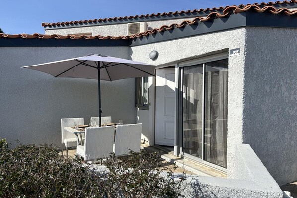 Une terrasse dotée de meubles en osier blanc et d’un grand parasol, d’une porte-fenêtre coulissante et d’un toit recouvert de tuiles.