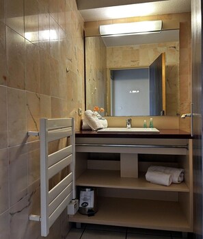 Un baño con espejo, lavamanos, perchero para toallas y un cubo de basura.
