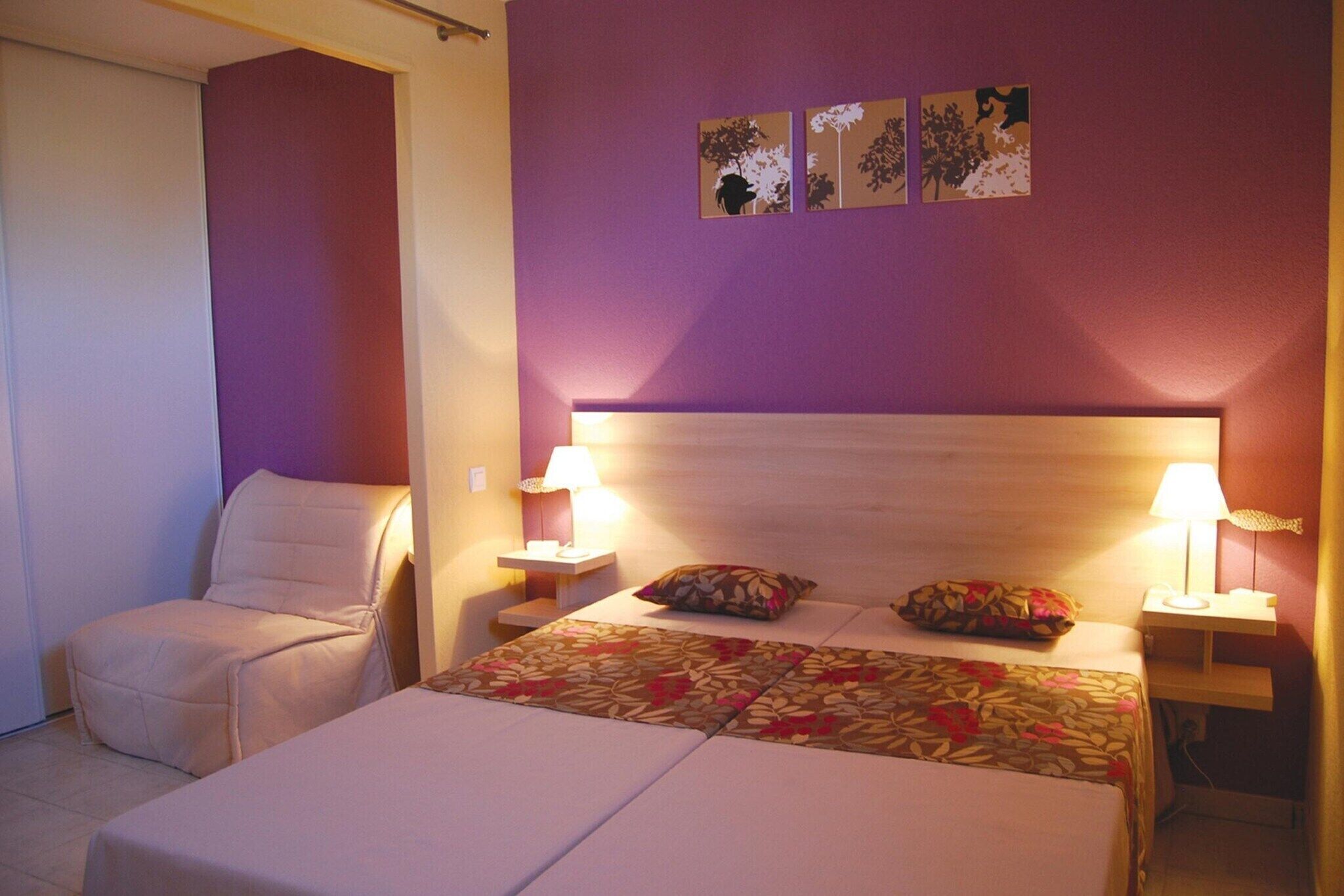 Una habitación de hotel con una cama, una silla, dos mesitas de noche con lámparas y una obra de arte en la pared.