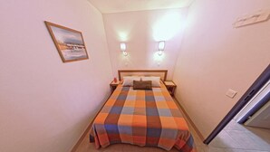Una habitaci ón de hotel pequeña con colcha a cuadros, dos lámparas de noche, una mesita de noche, un cuadro enmarcado en la pared y suelo de baldosas.