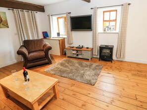 A living room with a brown leather armchair, a wooden coffee table with wine glasses, a TV, a fireplace, and a window with curtains. - The Hay Barn (Ripon)