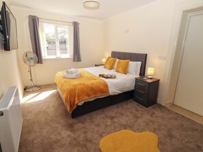 A hotel room with a bed, bedside table, lamp, and a standing fan. - Willow Cottage (Seahouses)