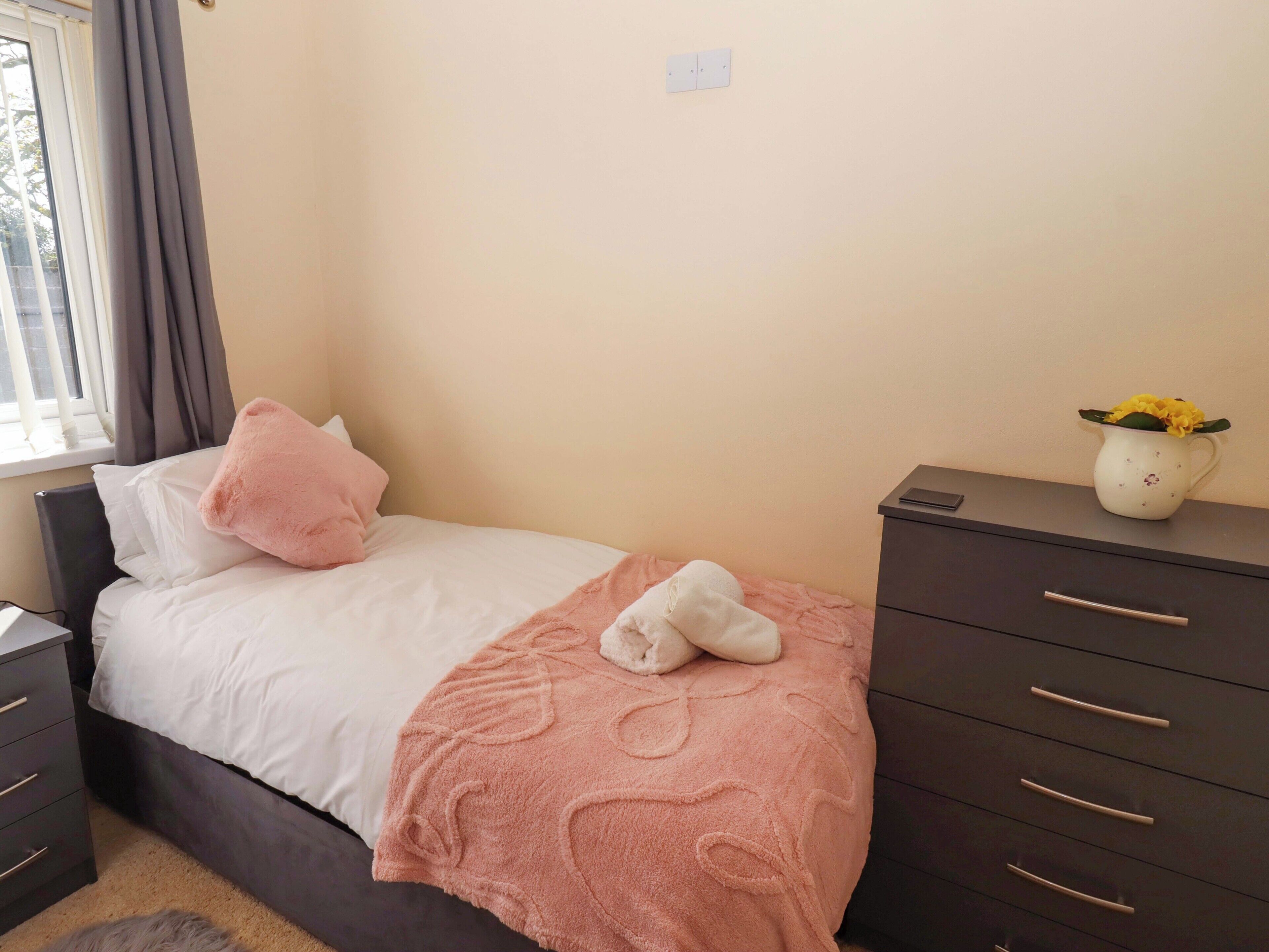 A bedroom with a bed, a bedside table, a dresser, a window with curtains, and a vase with flowers.