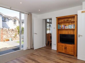 Ein Zimmer mit einem Holzboden-TV-Schrank, einer Tür, die auf eine Terrasse führt, und Blick auf ein Haus.