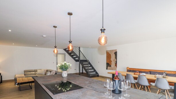 A modern kitchen with a central island, pendant lights, a staircase, and a dining area with chairs and a table. - Bigting (Rhosneigr)