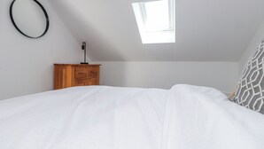 A bedroom with a bed, a wooden nightstand, a desk lamp, a window, and a circular wall decoration. - Bigting (Rhosneigr)
