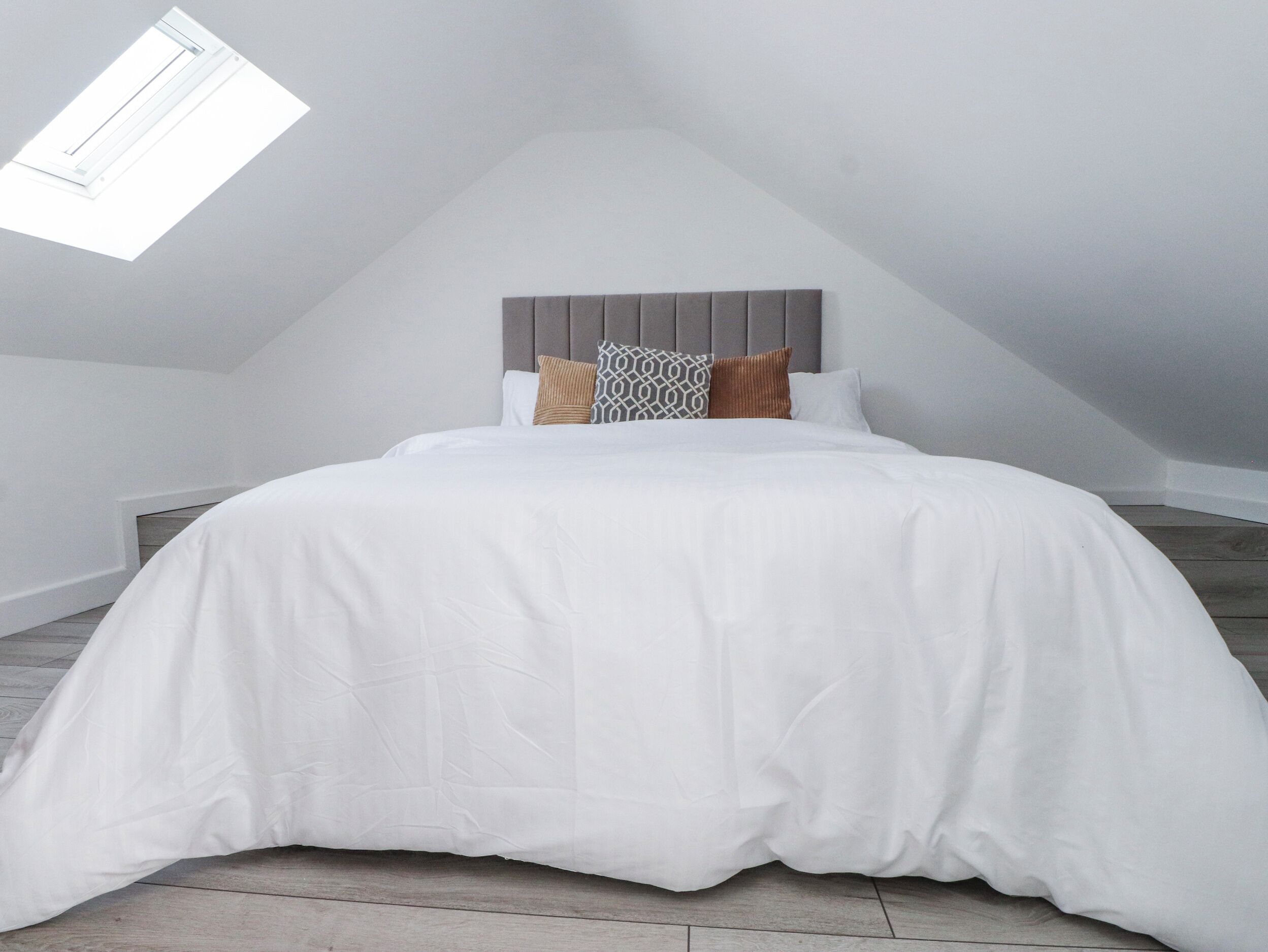 Un dormitorio con cama, cabecera, almohadas y tragaluz.