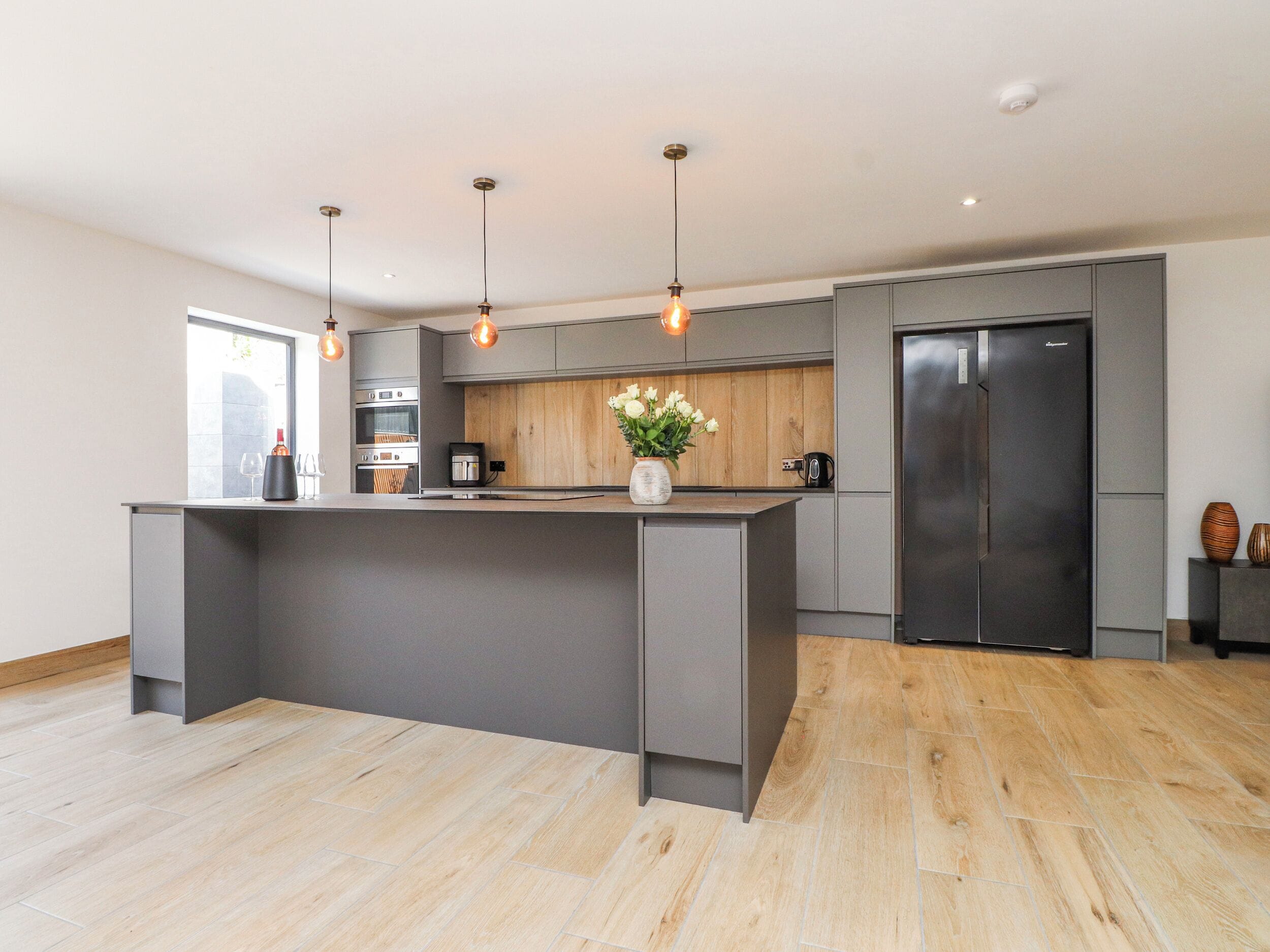 Een moderne keuken met een centraal eiland, inbouwapparatuur, hanglampen en een houten spatwand.