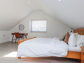 Un dormitorio con techo inclinado, cama con ropa de cama blanca, escritorio con silla y ventana con persianas.
