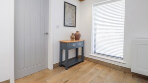 A hallway with a grey door, a small table with a vase, a framed artwork, and a window with blinds. - Bigting (Rhosneigr)
