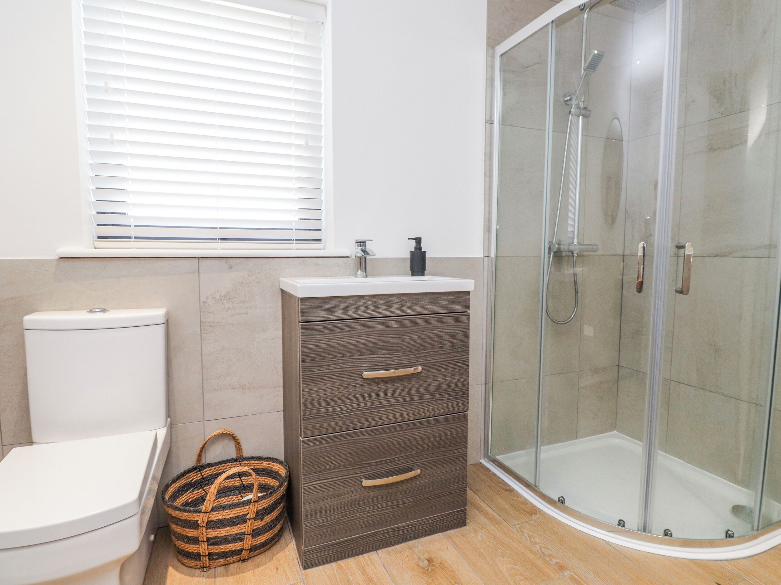 Un baño con inodoro, tocador con cajones, cabina de ducha de vidrio y ventana con persianas.