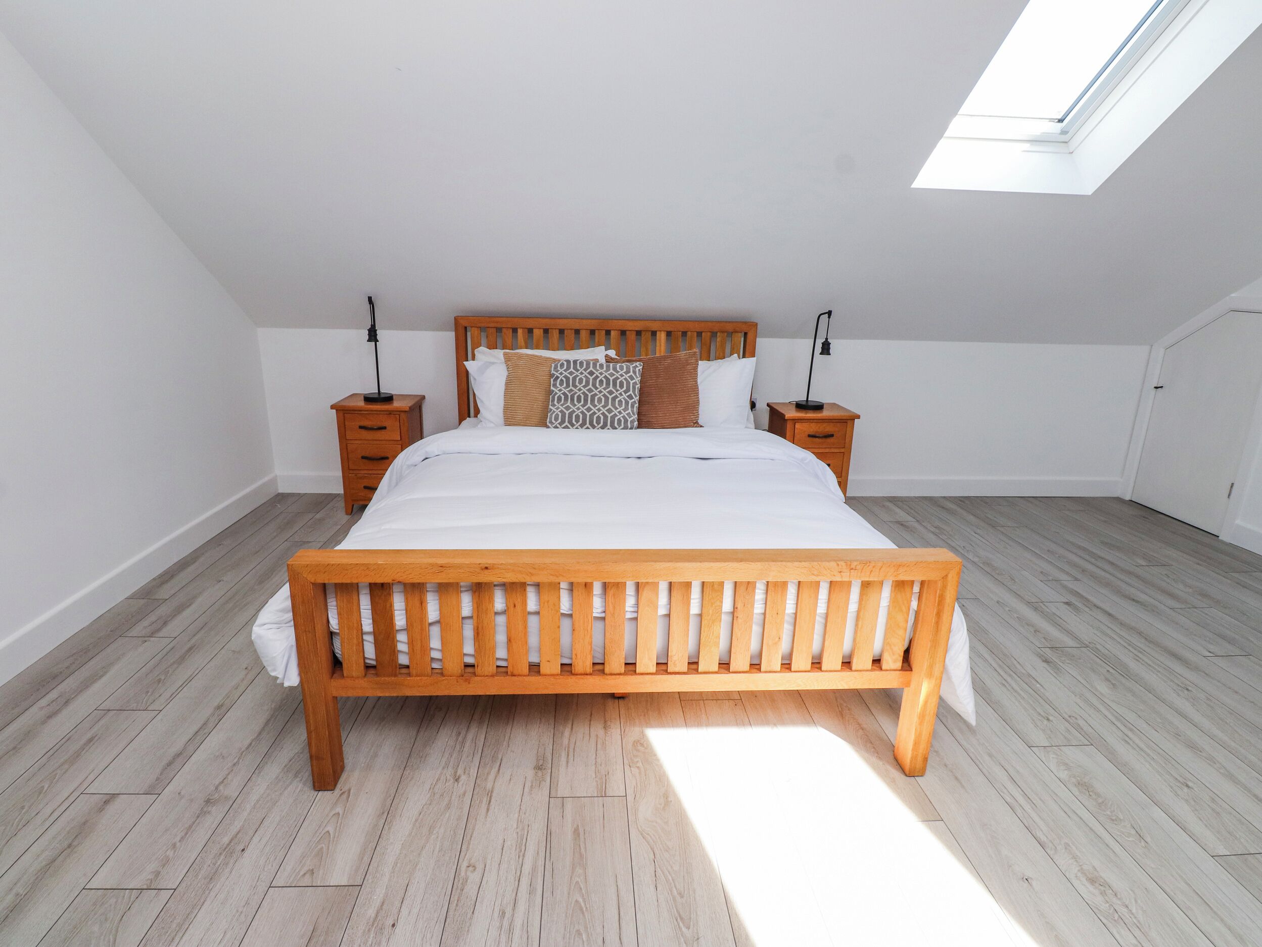 A bedroom with a wooden bed, two bedside tables, a skylight, and a view of the adjacent room.