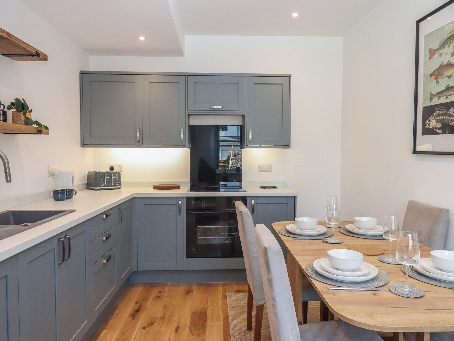 A modern kitchen with grey cabinets, a built-in oven, a dining table set for four, and a framed fish illustration on the wall.
