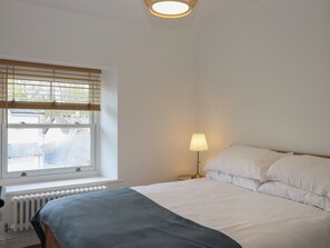 A bedroom with a bed, a window with blinds, a radiator, and a bedside lamp. - Hares Cottage (Torquay)