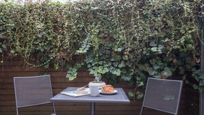Ein Tisch mit zwei Stühlen, ein Croissant, ein Buch und eine Tasse, vor einer mit Efeu bewachsenen Wand.