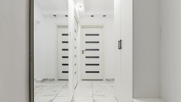 Um corredor com paredes brancas, chão de mármore e duas portas com fendas horizontais.