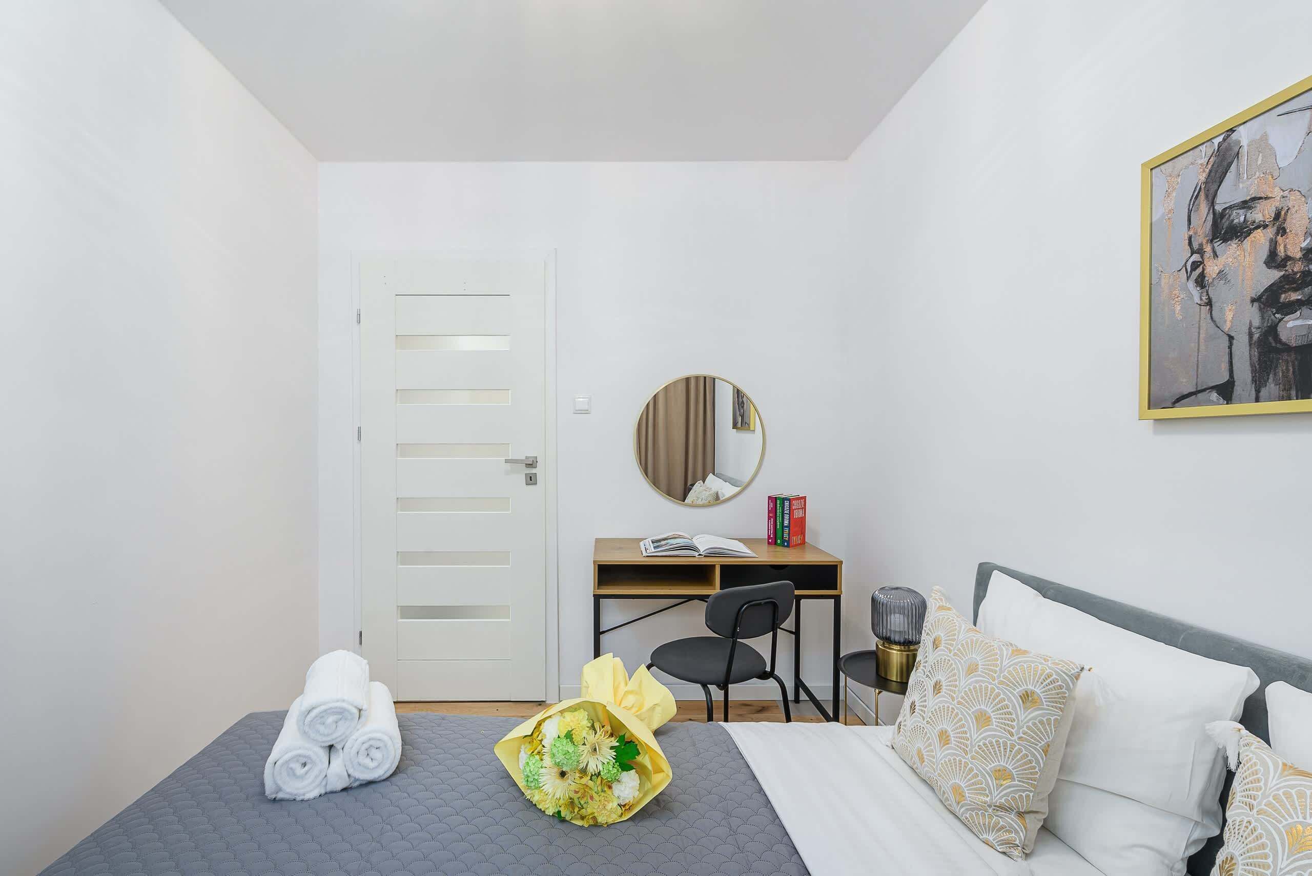 A hotel room with a bed, a desk, a chair, a round mirror, and a painting on the wall.
