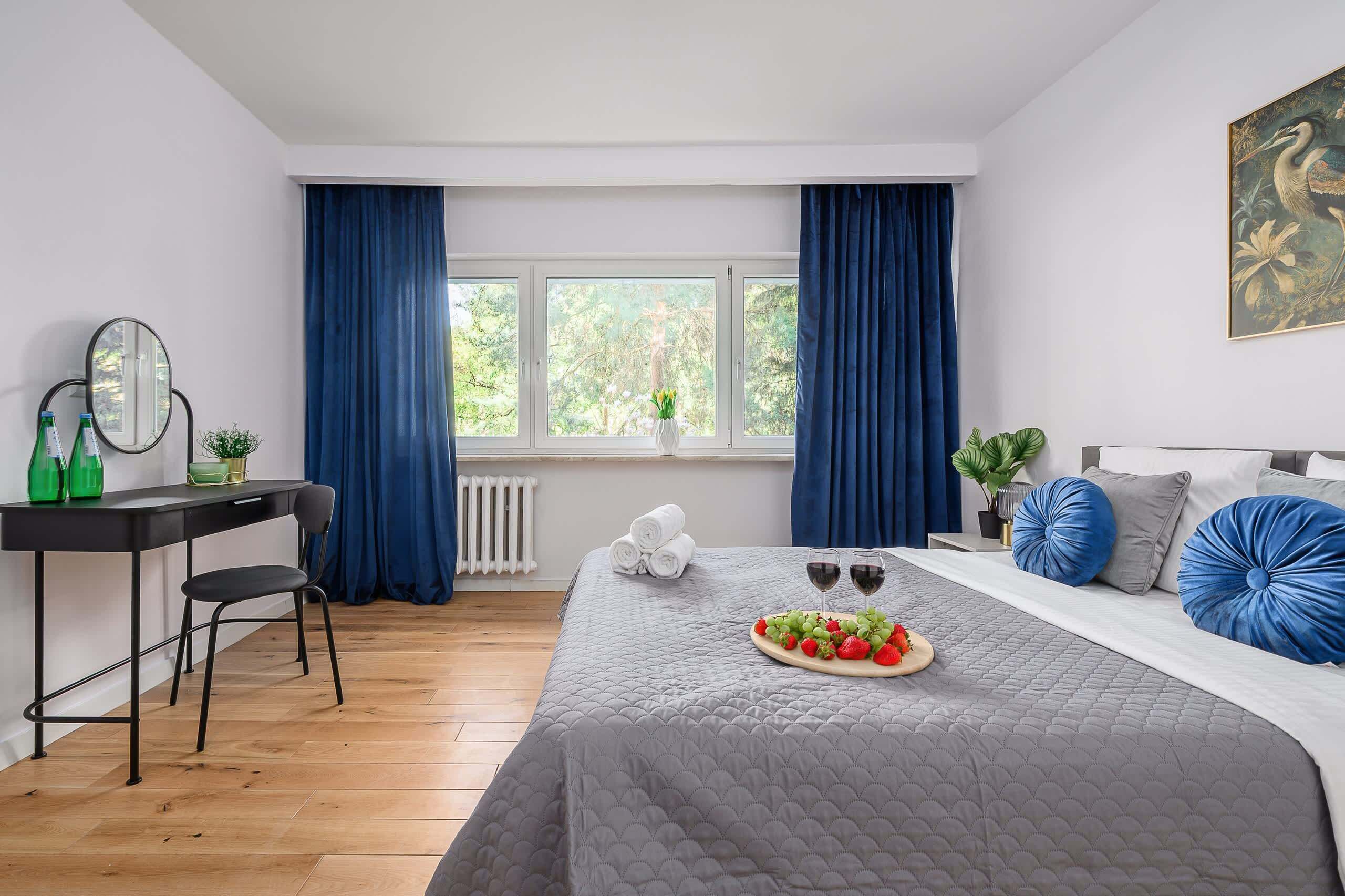 A hotel room with a bed, a desk, a chair, a radiator, a window with curtains, and a painting on the wall.