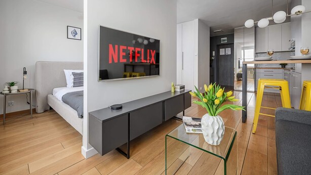 A modern living room with a bed, a black console table, a glass coffee table, a wall-mounted TV, and a kitchen area in the background. - Lumina apart near the Visla embankment (Warszawa)