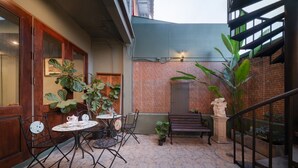 Un patio con pared de ladrillo, una puerta de madera, un banco de metal, una mesa con sillas y plantas en macetas.