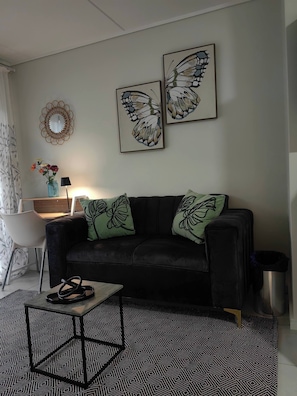 Una sala de estar con un sofá negro, dos obras de arte murales con forma de mariposa, un espejo redondo, una mesita con una lámpara y una alfombra estampada.