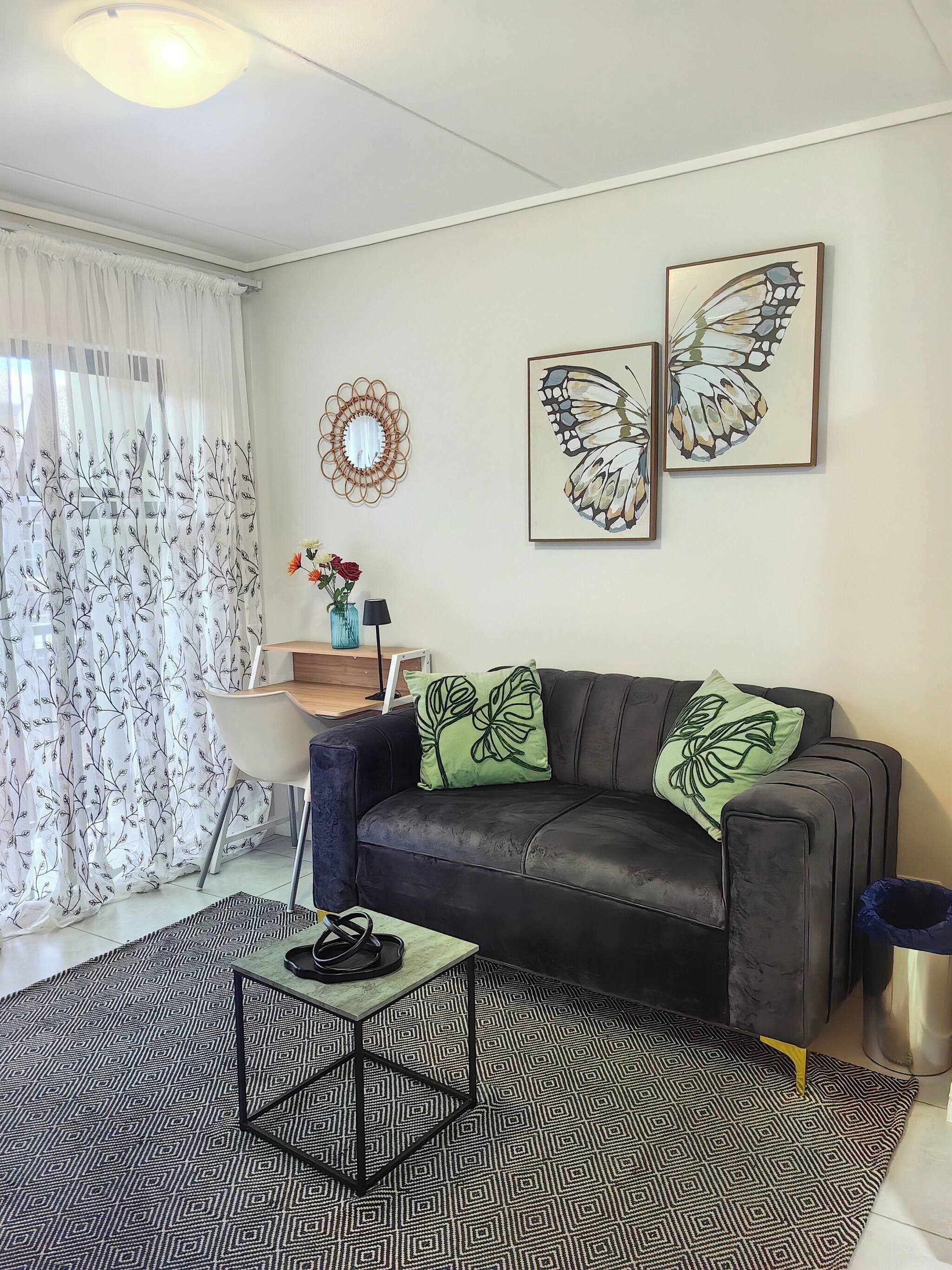 Uma sala de estar com sofá de couro preto, mesa de centro, tapete estampado, espelho fixado na parede e duas obras de arte com tema de borboletas.