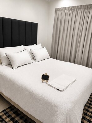 A neatly made bed with white bedding and pillows, a black headboard, and a small wooden easel with a blank canvas. - The Modern Sanctuary (Boksburg)