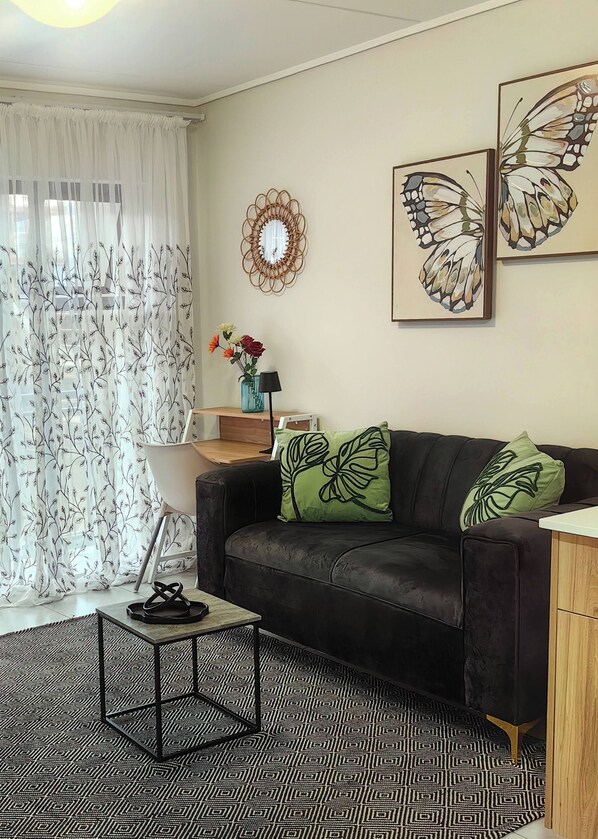 Una sala de estar con un sofá de cuero negro, cortinas estampadas, un espejo, arte con mariposas y un tapete con diseño.