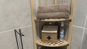 A corner bathroom with a wooden shelf unit containing a first aid kit, a bottle of air freshener, and a folded towel.