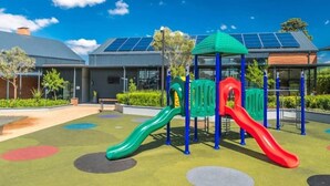 Un parque infantil con toboganes y una estructura para escalar, rodeado de un área ajardinada con un edificio que cuenta con paneles solares al fondo.