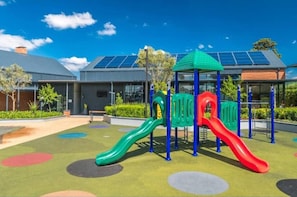 A playground with slides and a climbing structure, surrounded by a landscaped area with a building featuring solar panels in the background.