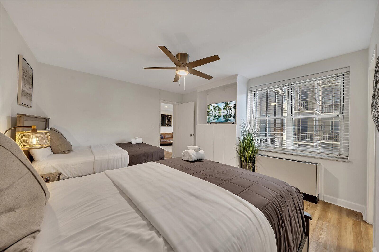 A hotel room with two beds, a ceiling fan, a window with blinds, and a plant.
