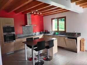 A modern kitchen with wooden cabinets, a central island with bar stools, a built-in oven, and a window with a view of greenery. - Family home in the mountains, in the Ossau Valley (Béost) (Béost)