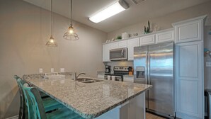 Una cucina con piano di lavoro in granito, elettrodomestici in acciaio inossidabile, sedie color petrolio e illuminazione a sospensione.