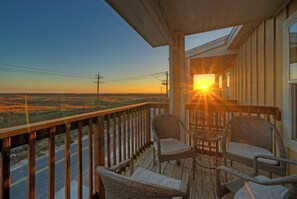A wooden deck with wicker chairs and a table, overlooking a road and fields during sunset. - Sleeps 32, 2 Homes | Heated Pool + Game Room + Balconies | Walk to the Beach (Corpus Christi)