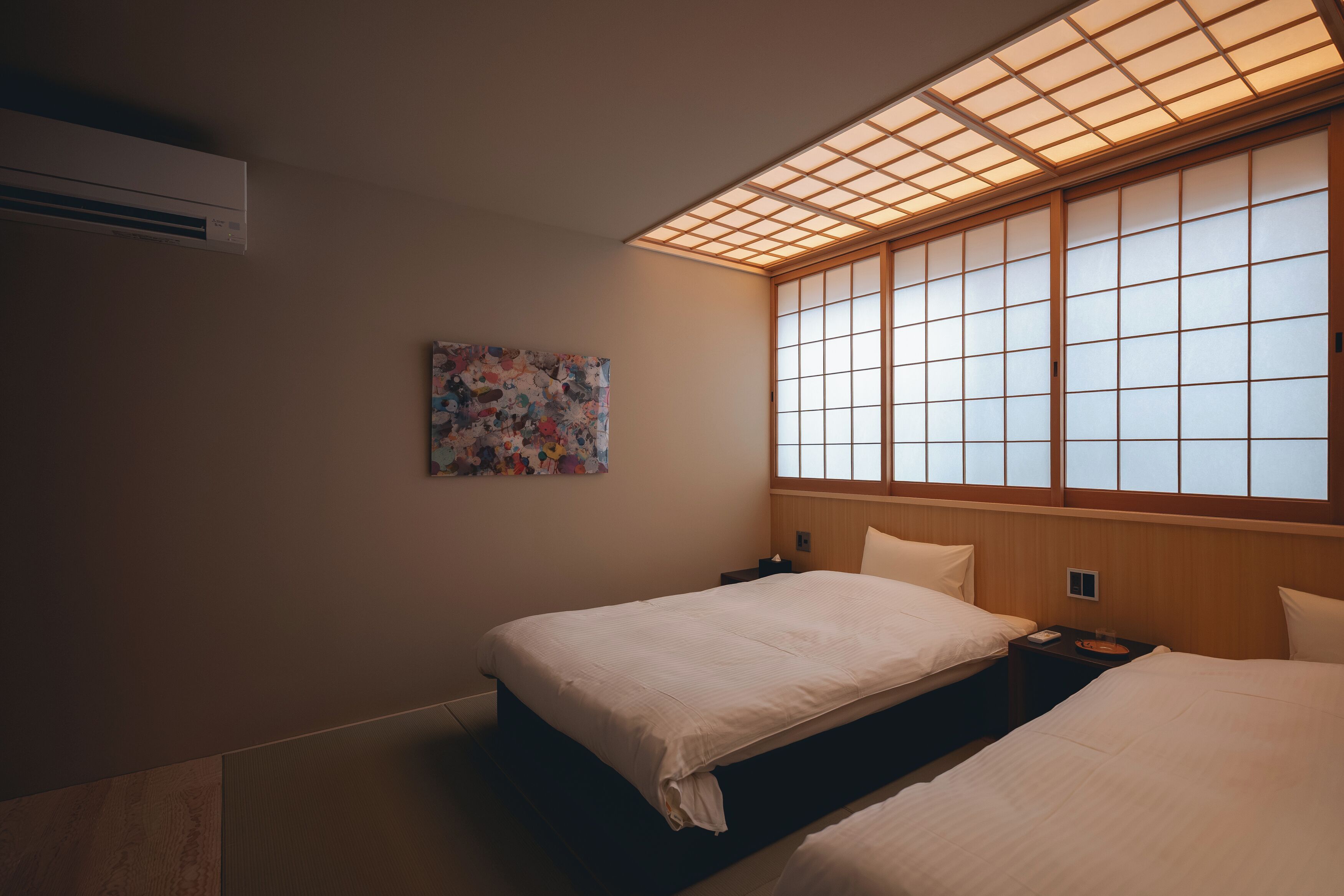 A hotel room with two beds, a large window with wooden frames, a wall-mounted air conditioner, and a colorful abstract painting.