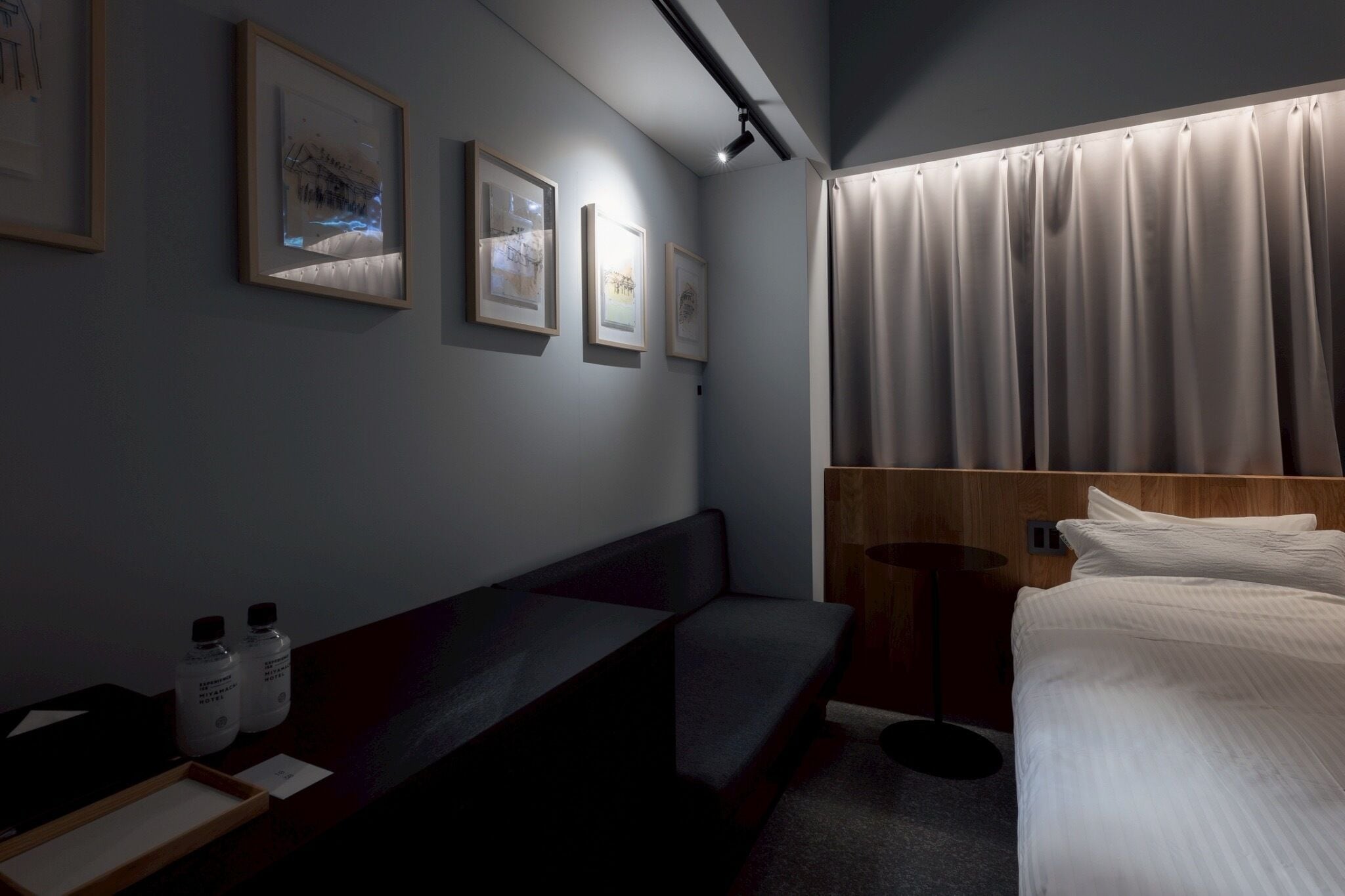 A hotel room with a bed, a bench, a bedside table with bottles, and framed pictures on the wall.